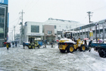 福生駅西口広場での重機による除雪作業のようすです。正面の白い建物は福生駅の自由通路で、昭和61年、橋上駅舎とともに完成しました。